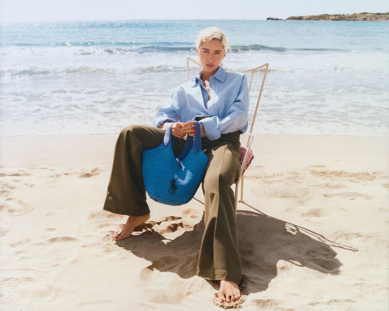 Woman in blue shirt and olive pants sits on beach chair, holding blue bag, with ocean and blue sky in background on sunny day.