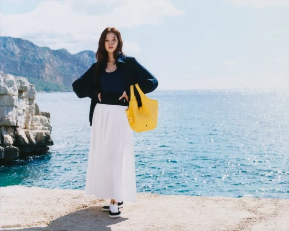 Woman in casual outfit with a yellow tote bag stands on a ledge overlooking the ocean, with cliffs and mountains in the background.