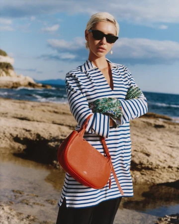 Model Iris Law wearing striped polo shirt and holding red leather bag by coastal rocks for Polo Ralph Lauren