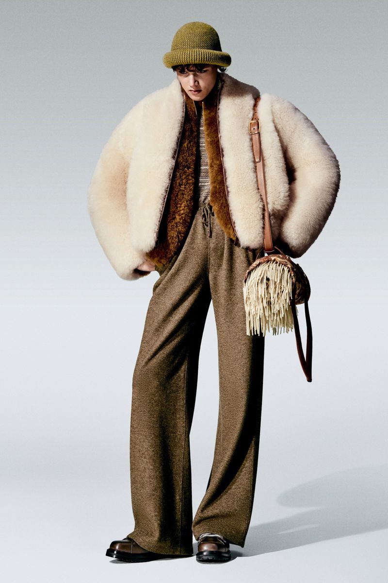 Fashionable person wearing a cream-colored fur coat, bucket hat, brown trousers, and a handbag with tassels in a studio setting.