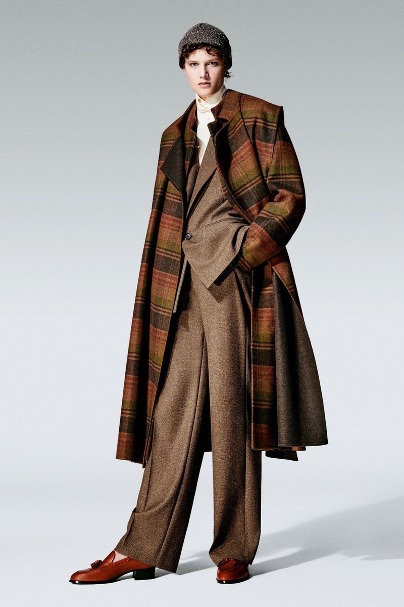 An adult fashion model is posing in a grey photo studio, wearing a brown suit, plaid overcoat, grey beanie, and brown leather loafers with tassels.