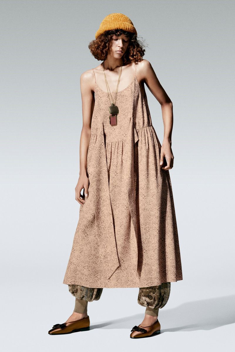 A female model wearing a loose, patterned dress, a yellow beanie, and flat shoes stands in a studio.