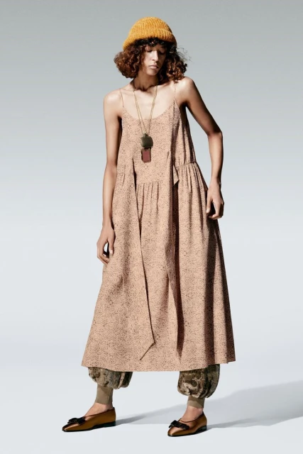 A female model wearing a loose, patterned dress, a yellow beanie, and flat shoes stands in a studio.