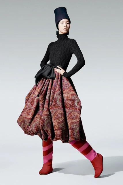 Fashion model poses in a black turtleneck, tall blue hat, full paisley skirt with a black bow, and red striped tights against a grey background.