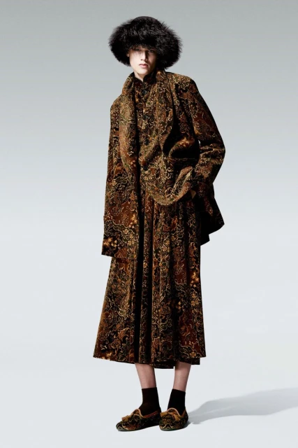 A single adult wearing a brown patterned jacket, skirt, and hat posing in a full shot against a white background in a studio.