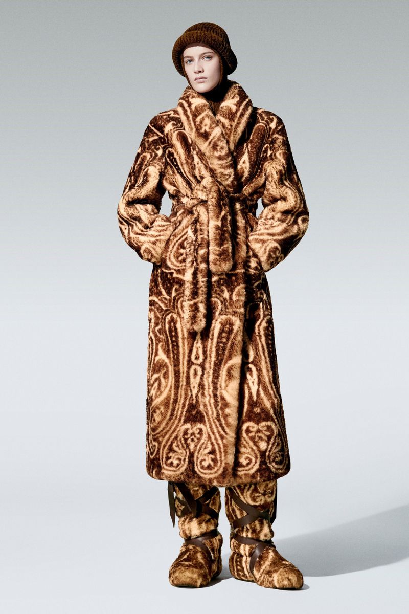 A model poses in an ornate brown fur coat, hat, and slippers against a light grey background in a studio fashion shot.