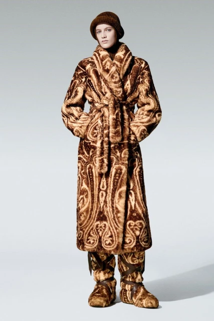 A model poses in an ornate brown fur coat, hat, and slippers against a light grey background in a studio fashion shot.