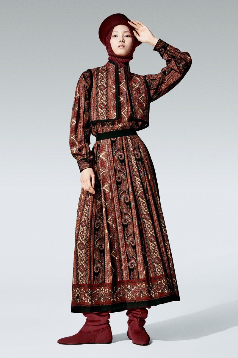 A fashion model poses in a long-sleeved, floor-length paisley dress with a matching cap, on a white background, wearing maroon sock boots.