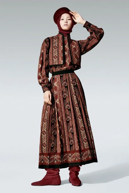 A fashion model poses in a long-sleeved, floor-length paisley dress with a matching cap, on a white background, wearing maroon sock boots.