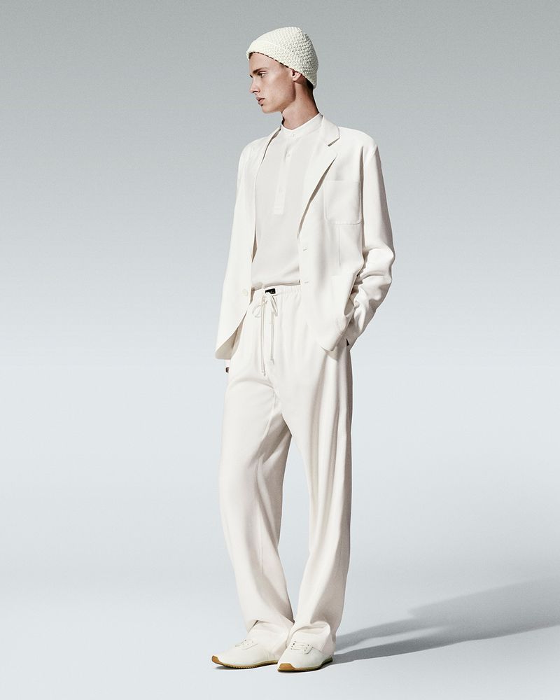 A young adult man wearing a cream-colored knit beanie, suit jacket, and slacks poses in a studio setting with a neutral gray background.