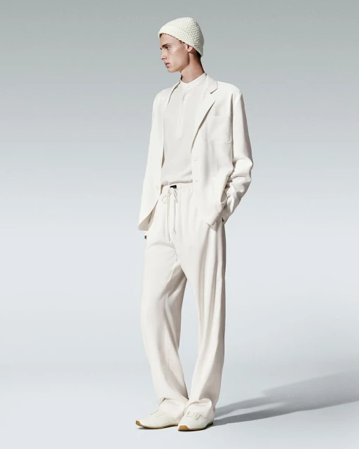 A young adult man wearing a cream-colored knit beanie, suit jacket, and slacks poses in a studio setting with a neutral gray background.