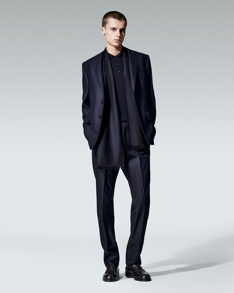 Fashionable adult man in a suit, shirt, and dress shoes posing in a full-body shot against a neutral grey background.