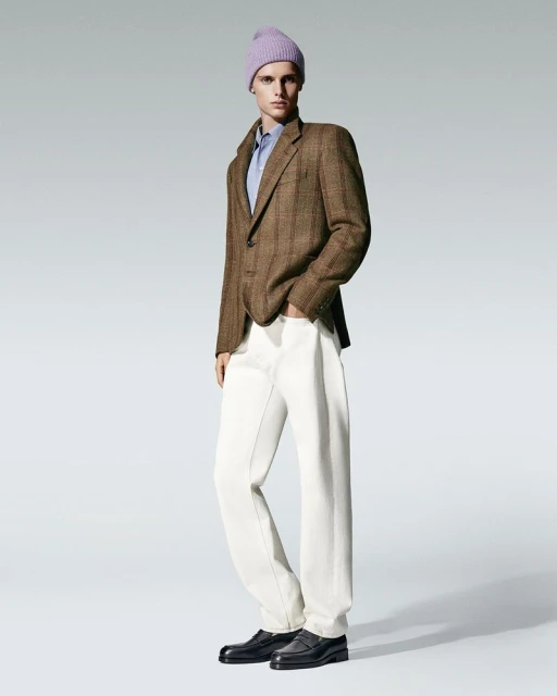 Fashionable man in a brown blazer, white pants, and lilac beanie poses in a studio with soft lighting, showcasing modern menswear.