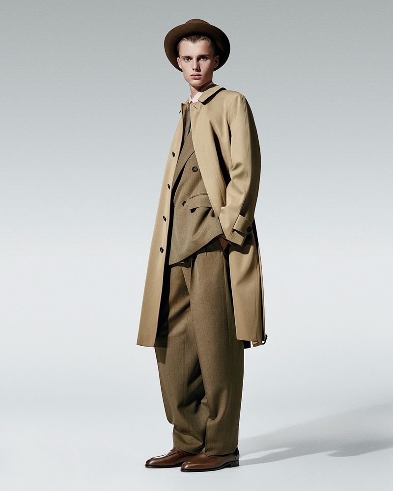 Model wearing a tan trench coat, brown suit, and hat against a light grey seamless studio background.