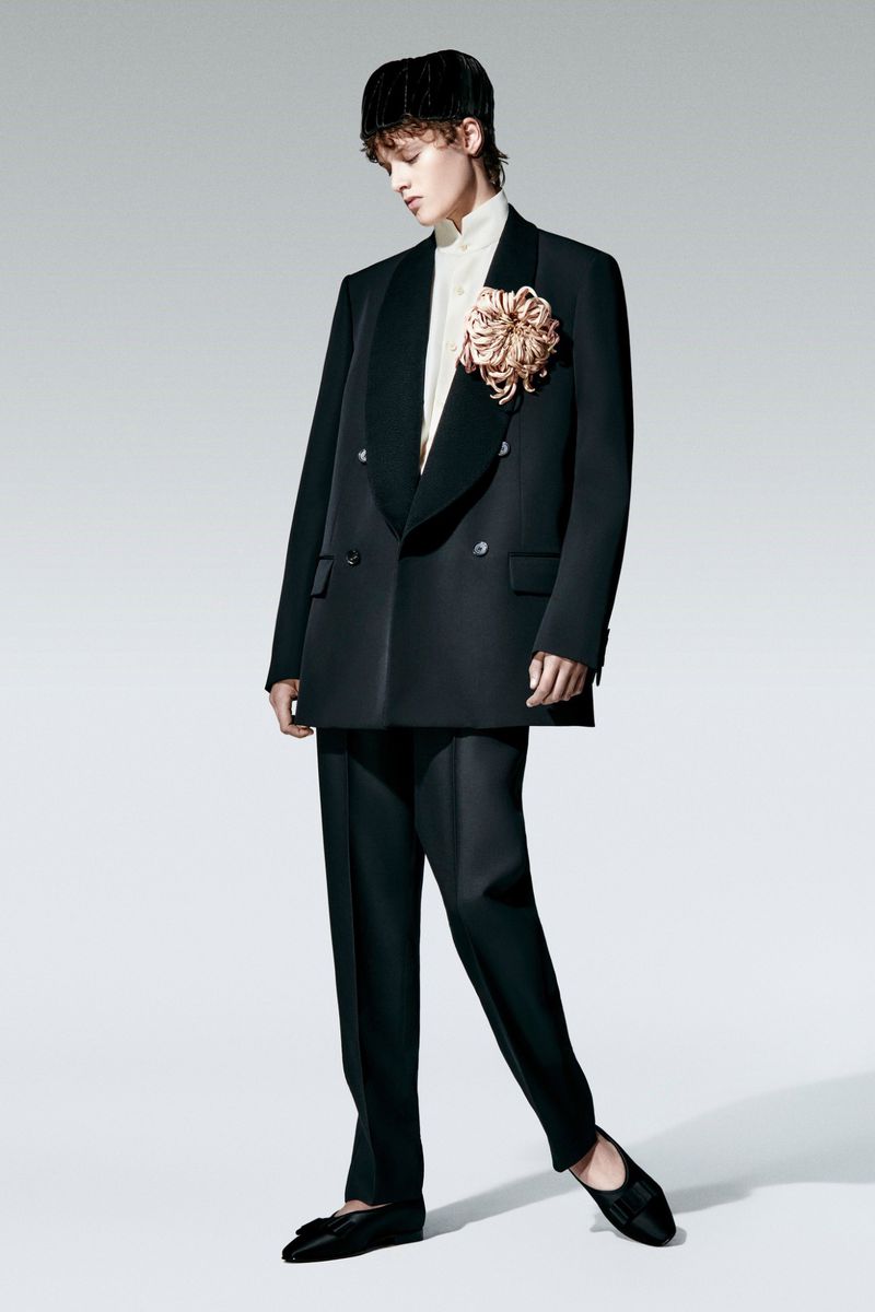 A person wearing a black double-breasted blazer with a large decorative flower, black trousers, and ballet flats poses in a studio.
