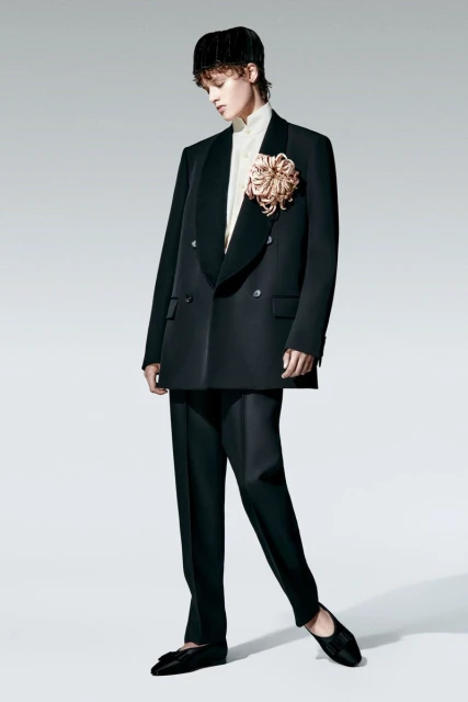 A person wearing a black double-breasted blazer with a large decorative flower, black trousers, and ballet flats poses in a studio.
