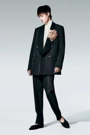 A person wearing a black double-breasted blazer with a large decorative flower, black trousers, and ballet flats poses in a studio.
