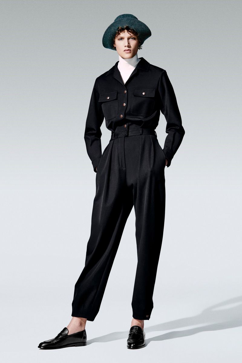 Full-length studio shot of a woman in a black button-down shirt, trousers, and a hat, posing against a gradient gray background.