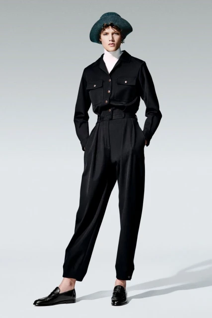 Full-length studio shot of a woman in a black button-down shirt, trousers, and a hat, posing against a gradient gray background.