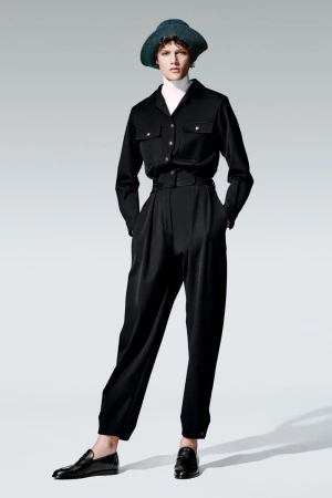 Full-length studio shot of a woman in a black button-down shirt, trousers, and a hat, posing against a gradient gray background.