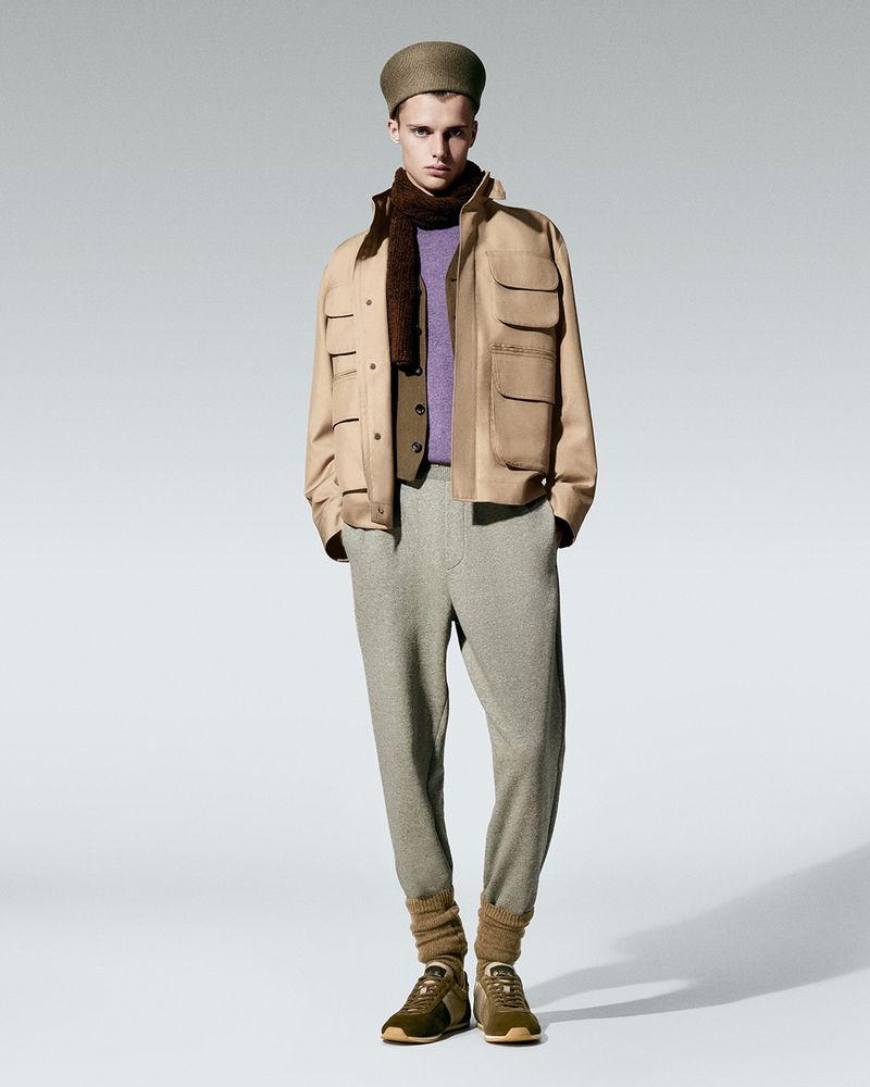 Full-length shot of a male model wearing a beige jacket, brown vest, purple sweater, grey sweatpants, leg warmers, and brown sneakers.