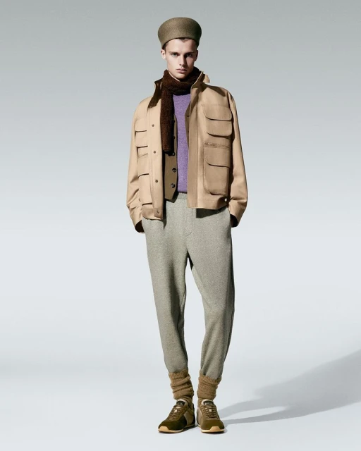 Full-length shot of a male model wearing a beige jacket, brown vest, purple sweater, grey sweatpants, leg warmers, and brown sneakers.