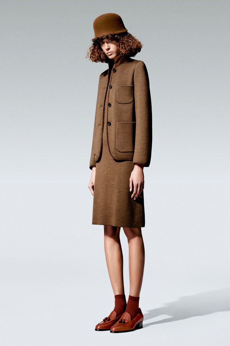 Woman poses in brown wool skirt suit, matching bucket hat, and red socks with brown loafers against a light gray background.