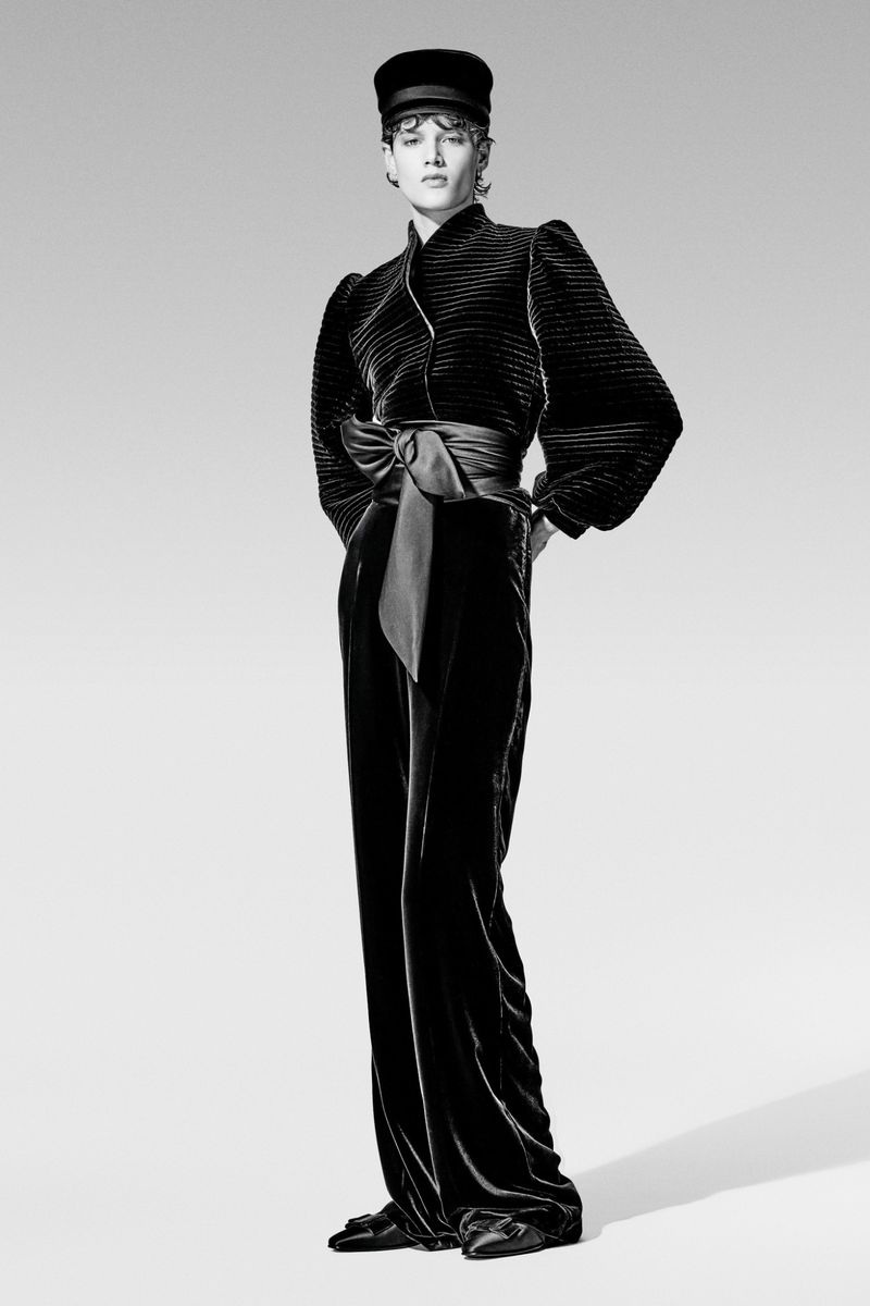 A black and white studio shot of a woman in an elegant black velvet outfit consisting of a top with a ribbon and wide pants.