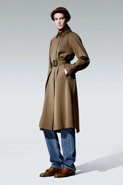 Woman in a brown trench coat, blue jeans, and brown hat and shoes, standing in a photo studio with neutral background.