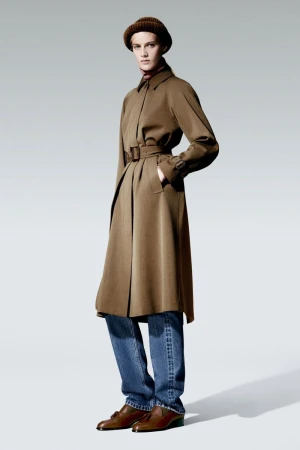 Woman in a brown trench coat, blue jeans, and brown hat and shoes, standing in a photo studio with neutral background.