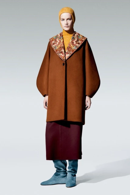 A woman in a brown coat with a patterned collar, a yellow turtleneck, and blue boots stands in a full shot against a grey backdrop.