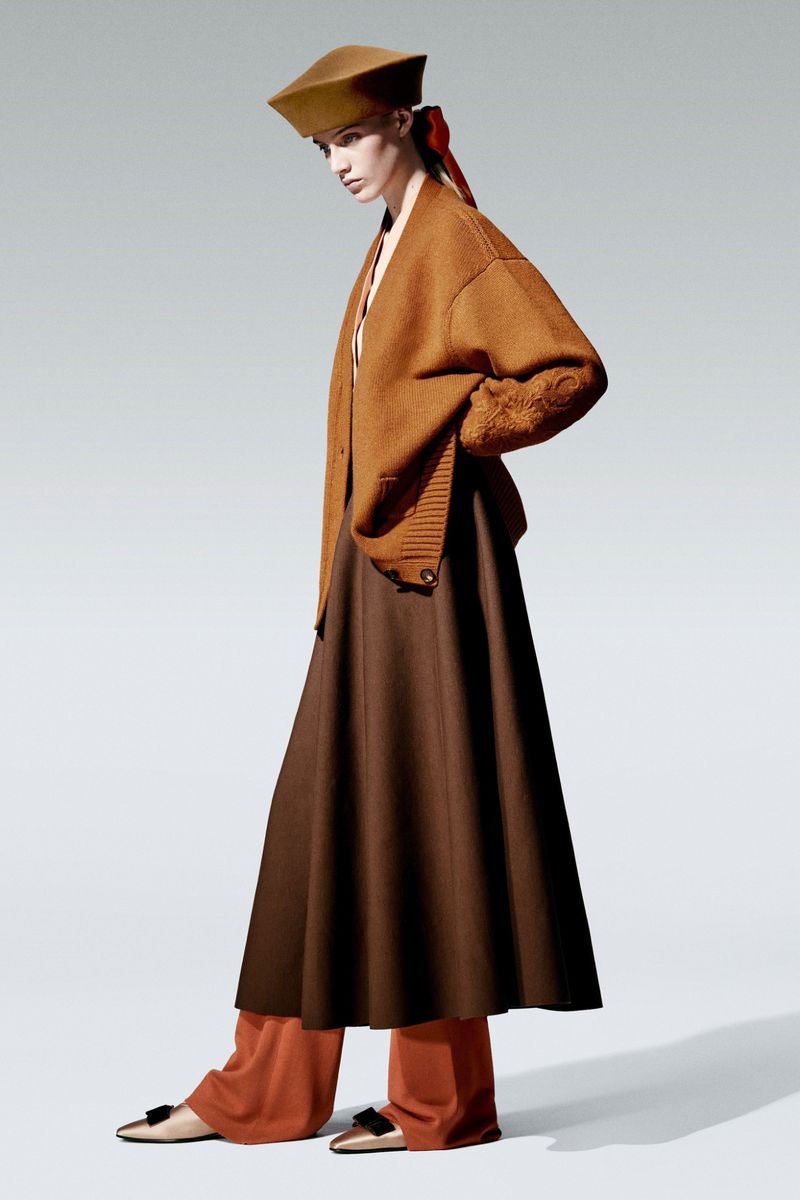 Adult female model wears a tan hat, caramel cardigan, dark brown skirt, and cream flats in a fashion editorial shot in a studio.