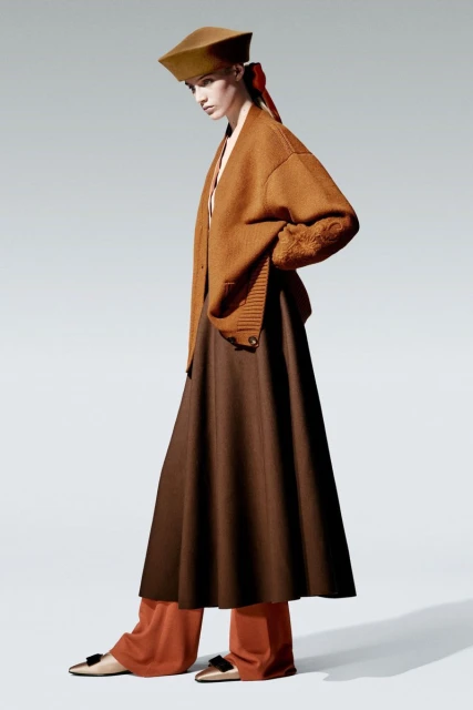 Adult female model wears a tan hat, caramel cardigan, dark brown skirt, and cream flats in a fashion editorial shot in a studio.