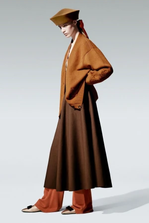 Adult female model wears a tan hat, caramel cardigan, dark brown skirt, and cream flats in a fashion editorial shot in a studio.