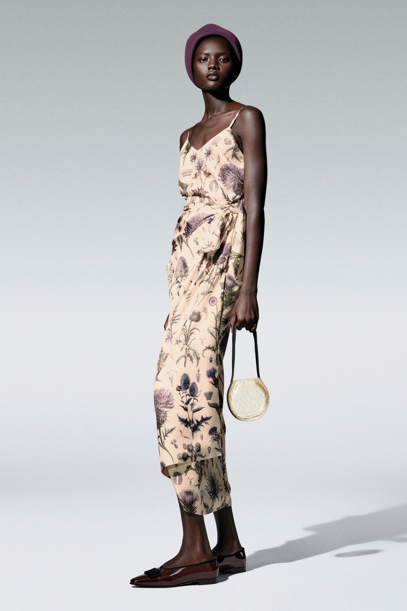 Full-length studio shot of an African woman in a floral-patterned dress, matching head covering, holding a round purse, and wearing flats.