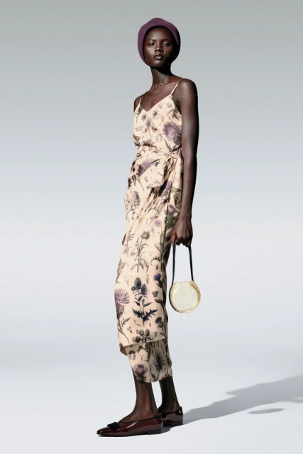 Full-length studio shot of an African woman in a floral-patterned dress, matching head covering, holding a round purse, and wearing flats.