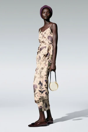 Model wearing floral wrap dress, dark purple beanie and round handbag standing against white studio backdrop