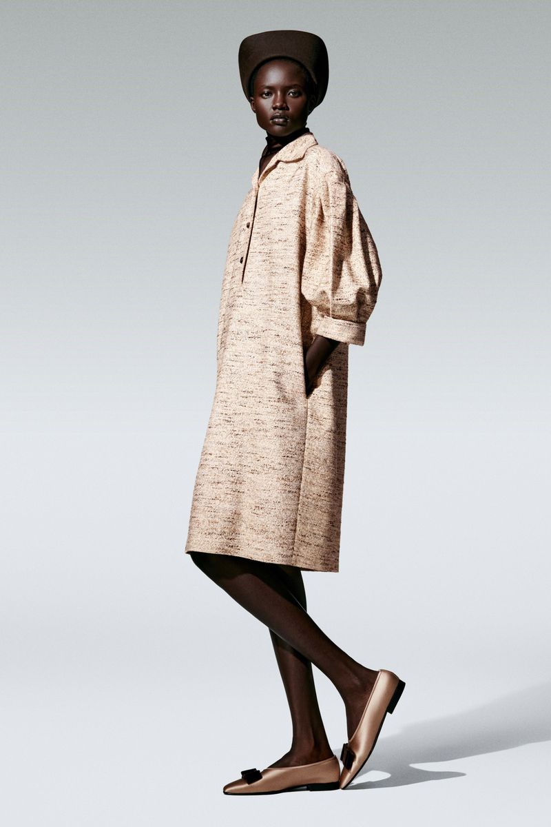 Fashion model posing in a studio, wearing a beige midi dress with balloon sleeves, a dark brown hat, and gold flats with black bows.