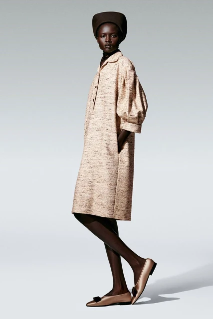 Fashion model posing in a studio, wearing a beige midi dress with balloon sleeves, a dark brown hat, and gold flats with black bows.