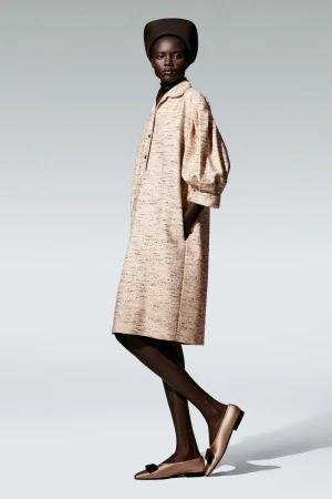 Fashion model posing in a studio, wearing a beige midi dress with balloon sleeves, a dark brown hat, and gold flats with black bows.