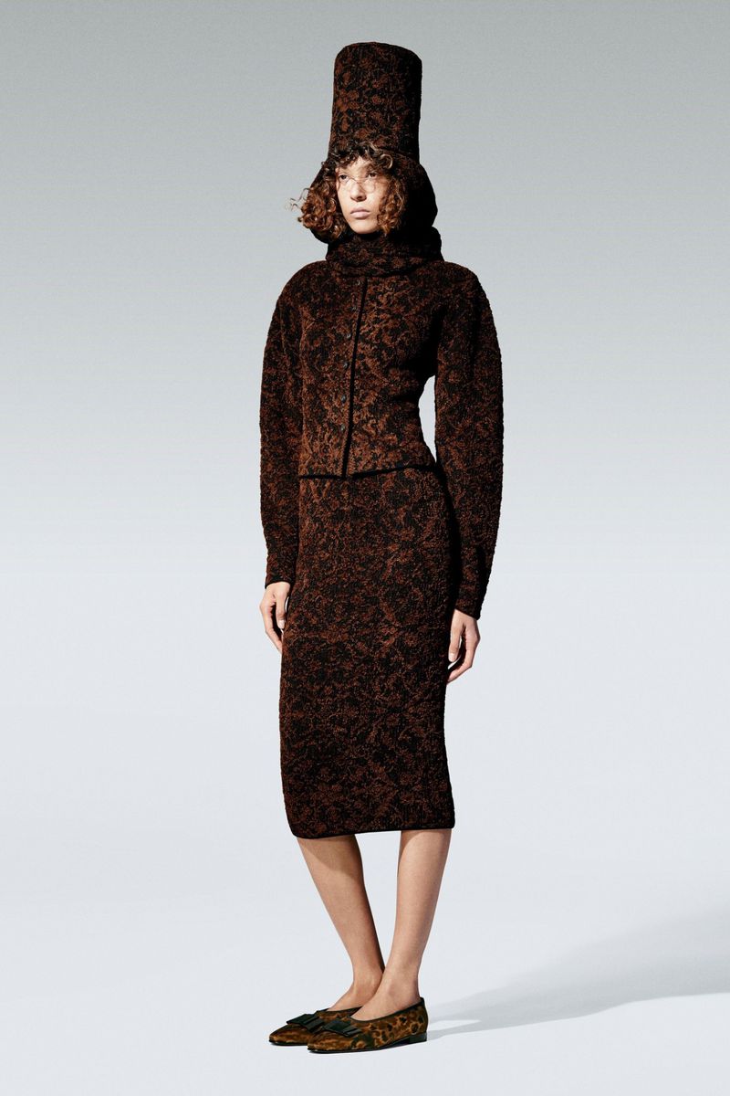Fashion model poses in a brown patterned knit outfit, including a high-neck top, skirt, tall cylindrical hat, and flats, on a white backdrop.