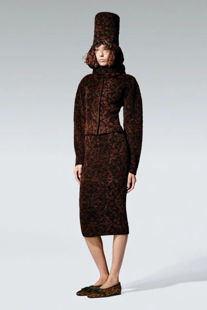 Fashion model poses in a brown patterned knit outfit, including a high-neck top, skirt, tall cylindrical hat, and flats, on a white backdrop.