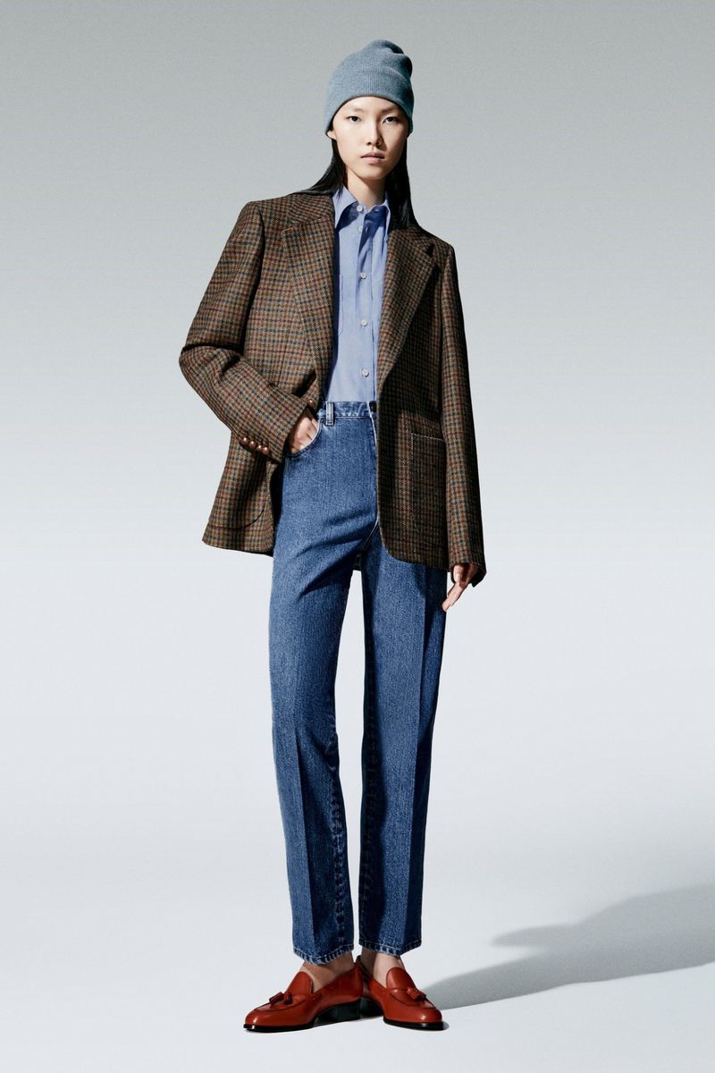 Studio shot of a young woman in a blue beanie, shirt, jeans and brown blazer, standing on a white background wearing brown loafers.
