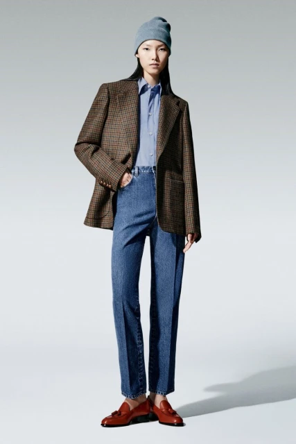Studio shot of a young woman in a blue beanie, shirt, jeans and brown blazer, standing on a white background wearing brown loafers.