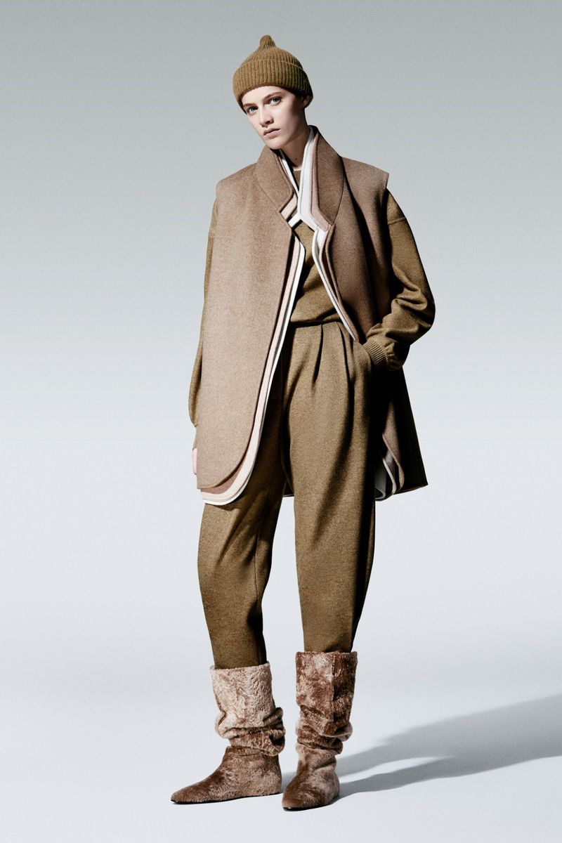 A woman in a tan outfit poses in a studio setting, showcasing fashion with a beanie, long vest, sweater, wide-leg pants, and distinctive boots.