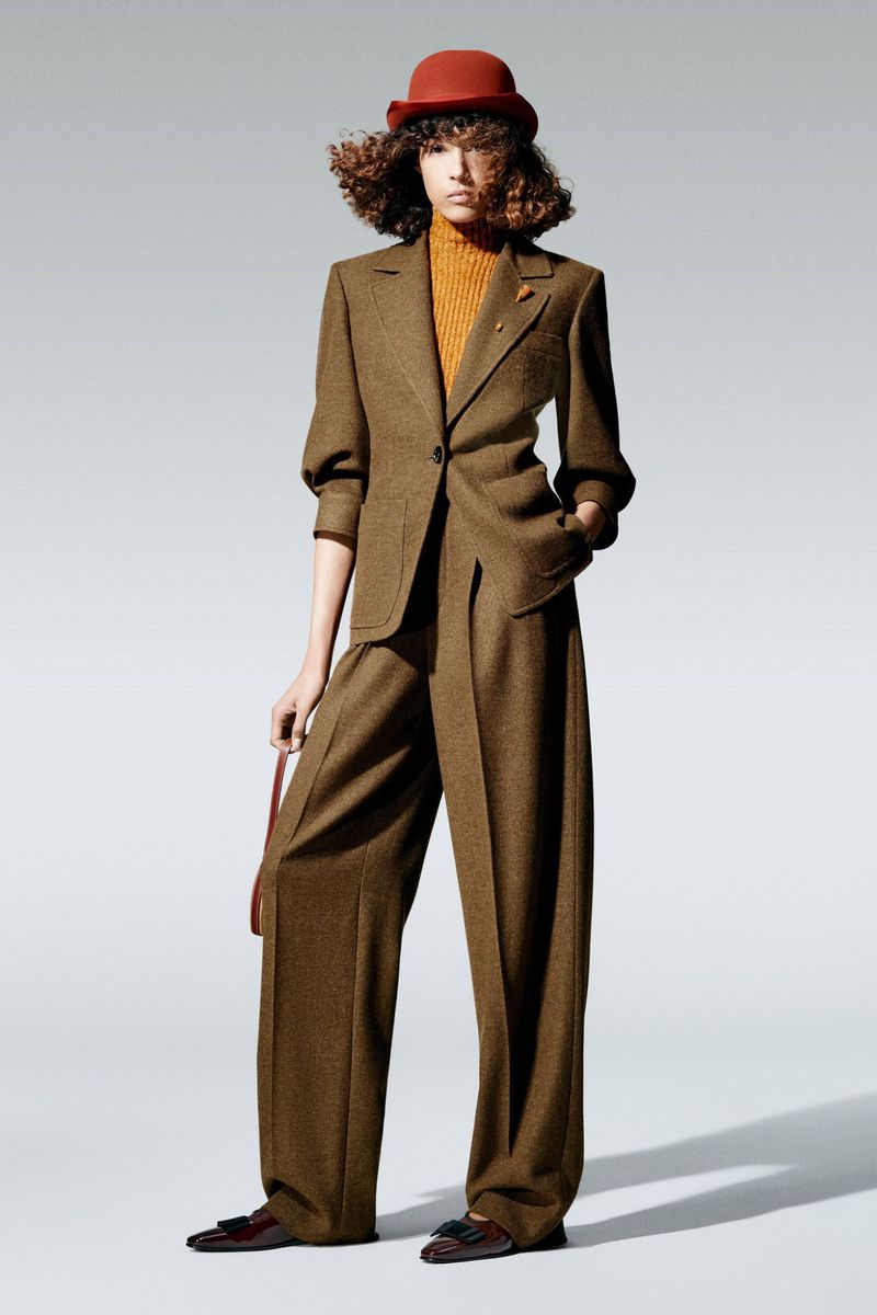 A woman poses in a brown tweed suit with a bowler hat, standing in a studio with a neutral background, showcasing high fashion.