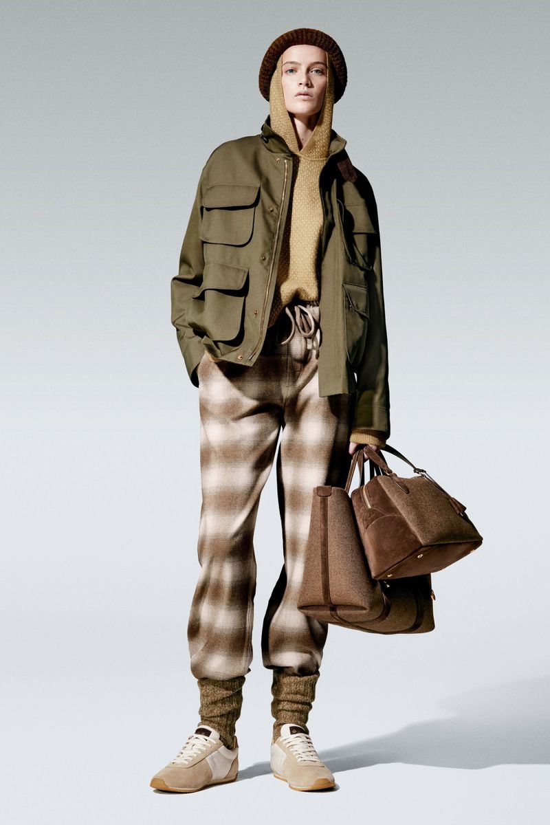 Woman in olive jacket, beige sweater, plaid pants, beanie, holding purses. Fashion shot on a light background in studio lighting.