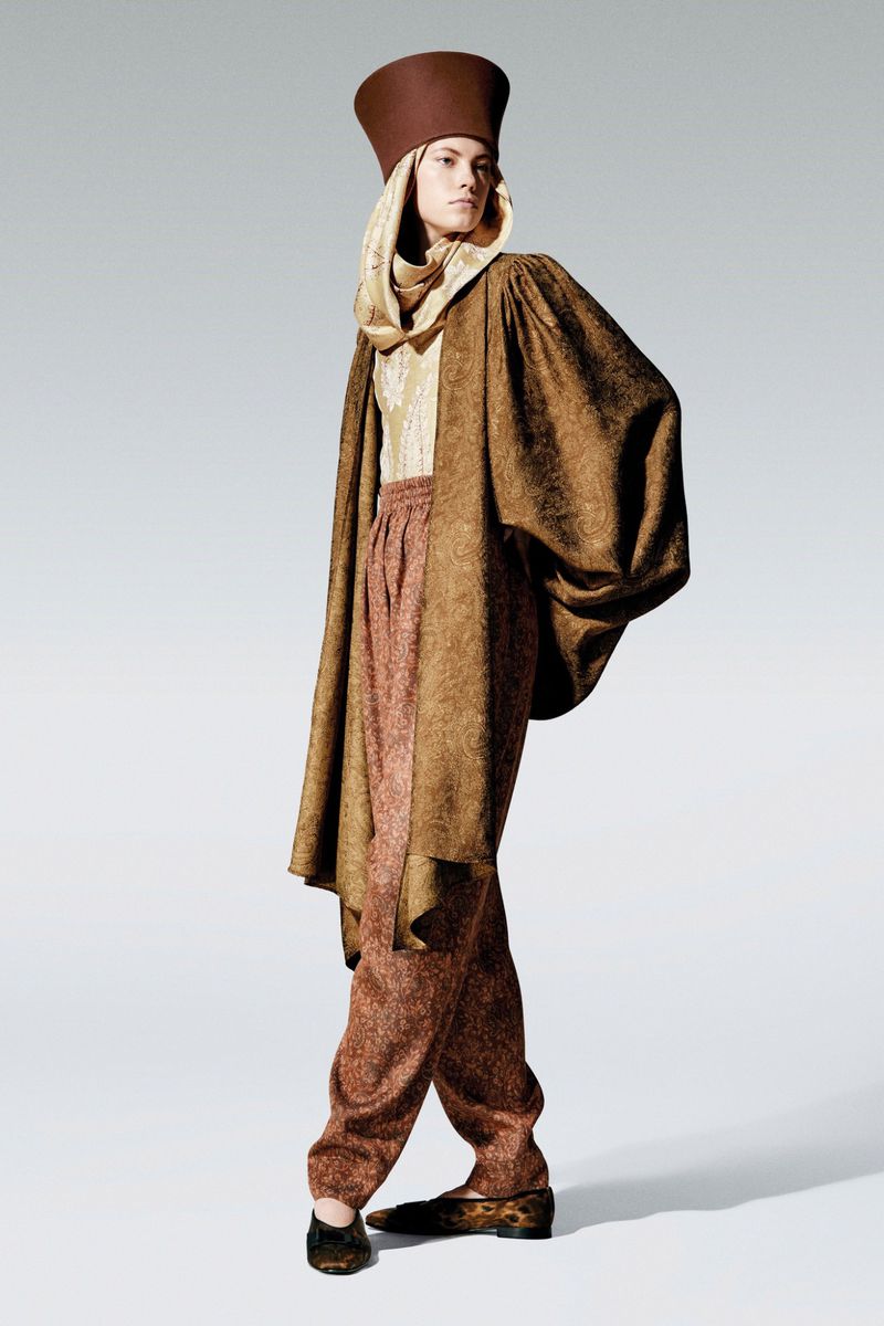 Full-length fashion shot of a woman in brown textured clothing and a tall hat against a light gray backdrop in a photo studio.