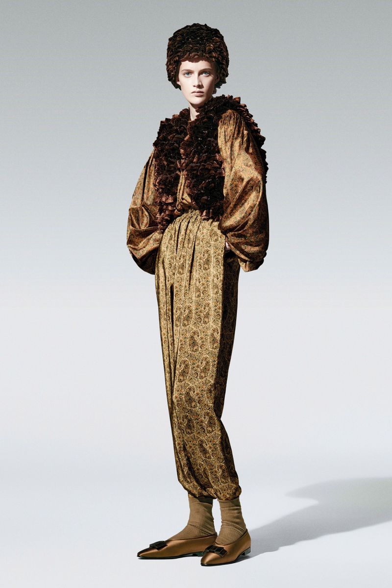 Fashion model stands in studio wearing a golden paisley patterned blouse and trousers with a brown ruffled jacket and hat ensemble.