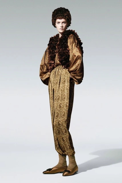 Fashion model stands in studio wearing a golden paisley patterned blouse and trousers with a brown ruffled jacket and hat ensemble.